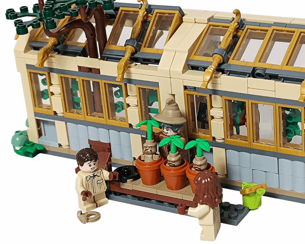 LEGO Harry Potter 76445 ホグワーツ城：薬草学の授業 マンドレイクの授業シーン。 スプラウト先生が中央でマンドレイクを掲げ、両側にハーマイオニーとネビルが立つ。 温室の外壁と木の幹、オレンジ色の屋根窓が背景に映り、ジョウロと鉢が並んでいる。