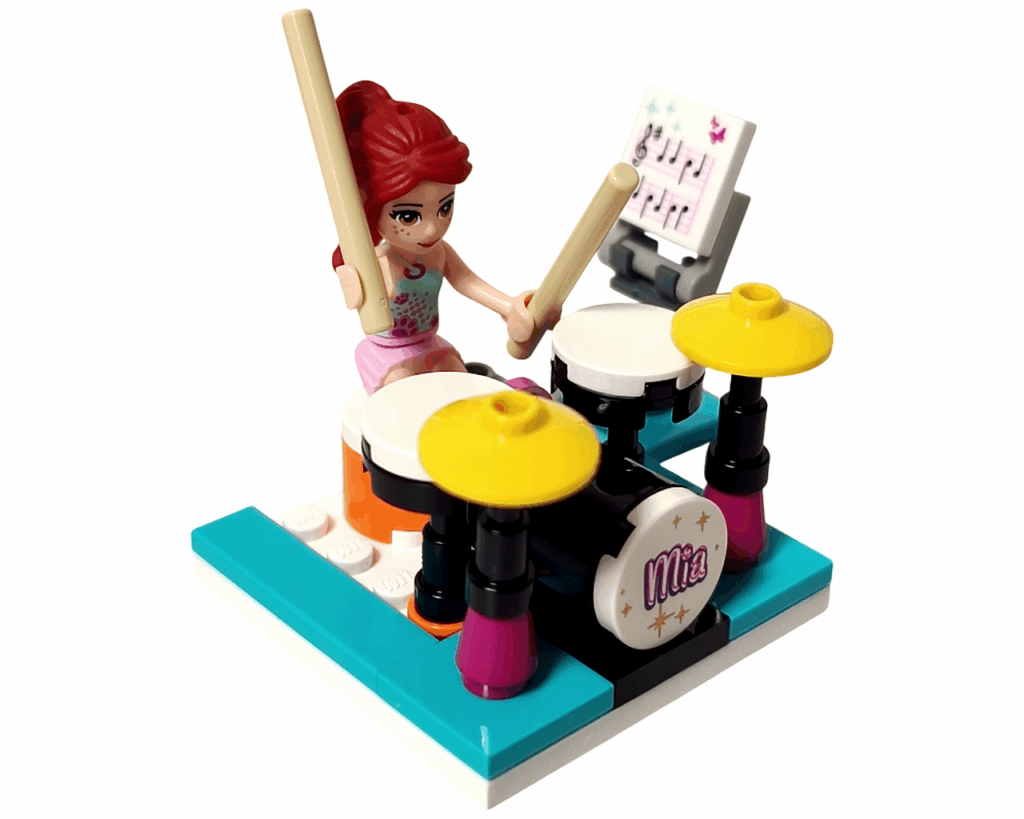 ミニドールのミアがスティックを持ち、黄色いシンバル付きのドラムセットを叩いている。後ろには譜面台がある